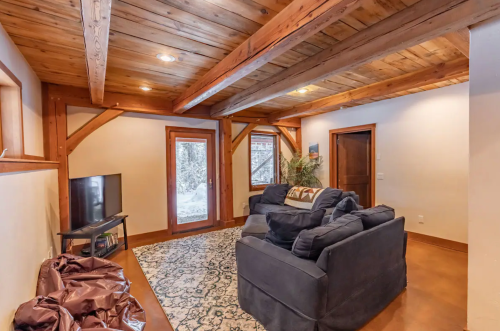 Cozy living room with wooden beams, a dark sofa, a TV, and large windows overlooking a snowy landscape.